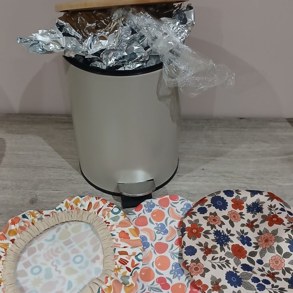 Poubelle remplie de film plastique et de papier aluminium à côté de plusieurs charlottes alimentaires réutilisables en tissu, illustrant une alternative durable pour conserver les aliments.